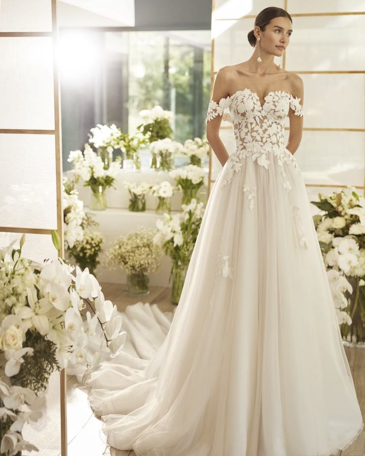 In questa foto una modella indossa un abito ad con ricami di pizzo floreale ramage che dal corpetto dallo scollo a cuore off shoulder scende sulla gonna in tulle. Il modello fa parte della collezione sposa Rosa Clará Couture 2024