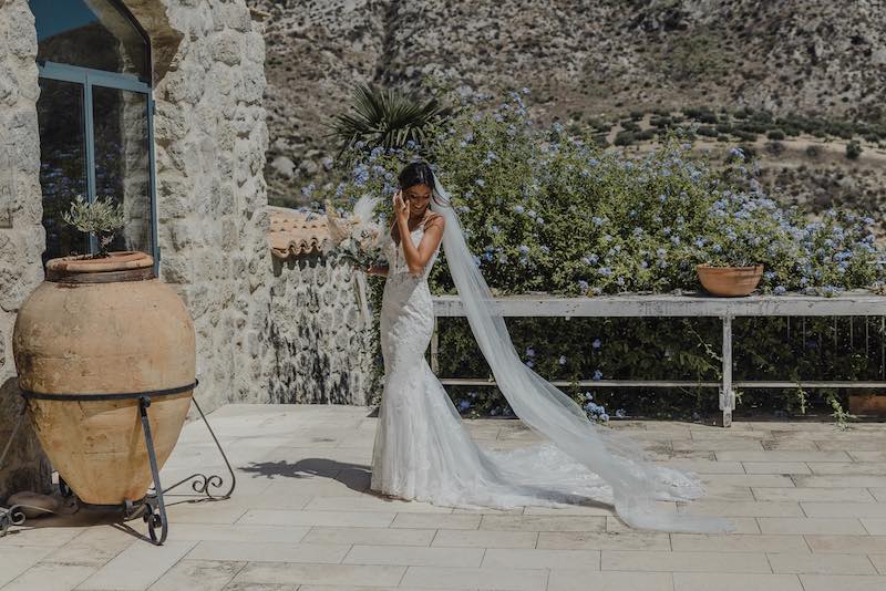In questa foto di Alessandro Tondo una sposa sorride su una terrazza circondata da fiori mentre guarda in basso, si aggiusta i capelli con la mano sinistra mentre con la mano destra tiene in mano il suo bouquet. La sposa indossa un abito a mezza sirena in pizzo e un velo di tulle taglio laser