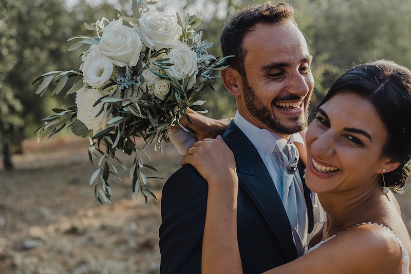 In questa foto di Alessandro Tondo due sposi si abbracciano e sorridono alla macchina fotografica. La sposa mostra il bouquet di fiori di colori bianchi