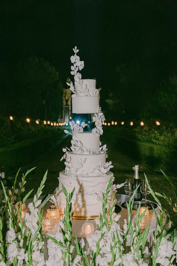 In questa foto la torta monumentale del matrimonio di Carlotta e Federico di colore bianco, a piani e con foglie e fiori 3D attorno alla quale sono presenti decorazioni di fiori di colore bianco e piccole candele 