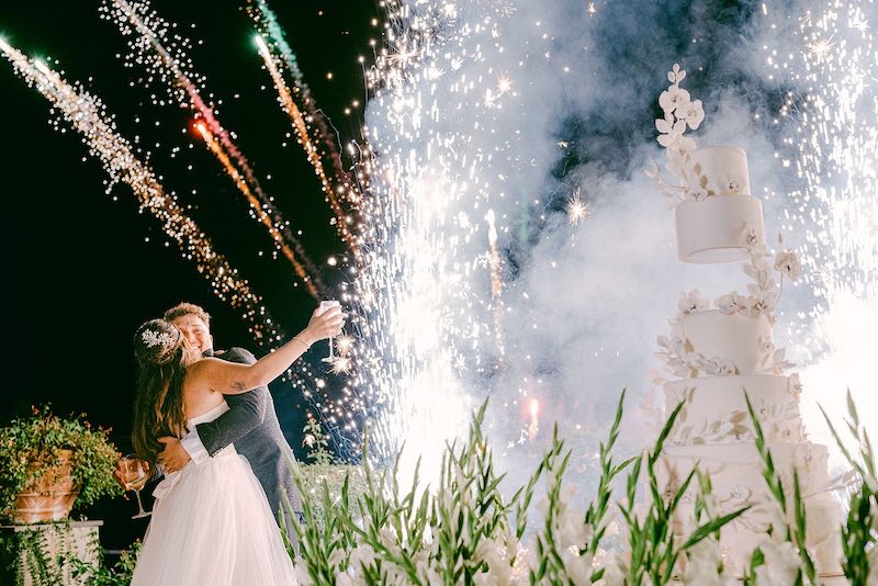 In questa foto gli sposi Carlotta e Federico si abbracciano dietro alla loro torta nuziale di colore bianco e a piani circondata da fiori di colore bianco e da candele dall'esplosione di fuochi d'artificio