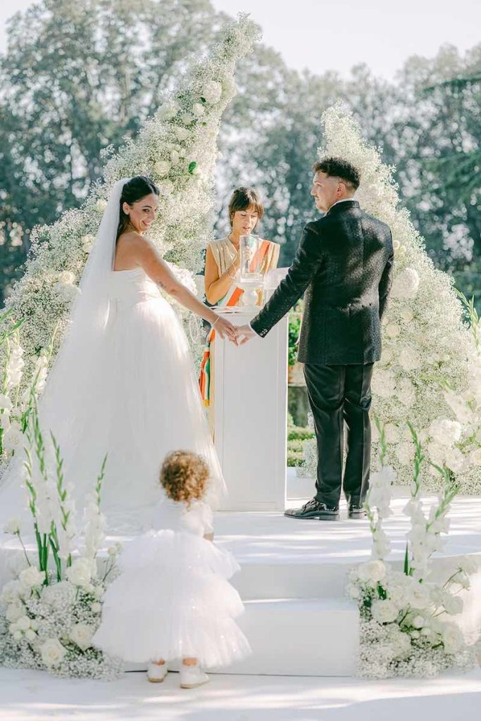 In questa foto un momento del matrimonio civile di Carlotta e Federico davanti ad un altare di fiori di colore bianco. Gli sposi sono leggermente girati di spalle per guardare la loro bimba mentre sale le scale indossando un abito con gonna di tulle