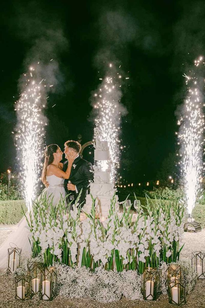 In questa foto gli sposi Carlotta e Federico si baciano dietro alla loro torta nuziale di colore bianco e a piani circondata da fiori di colore bianco e da candele dall'esplosione di fuochi d'artificio