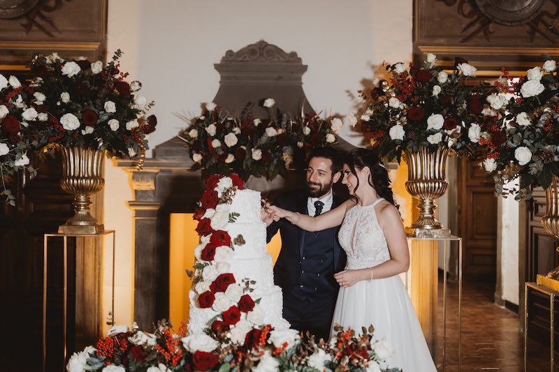 In questa foto Matteo e Pina taglia la loro torta nuziale del matrimonio di Matteo e Pina, a piani decorata con crema al burro di colore bianco e fiori freschi di colore rosso e bianco