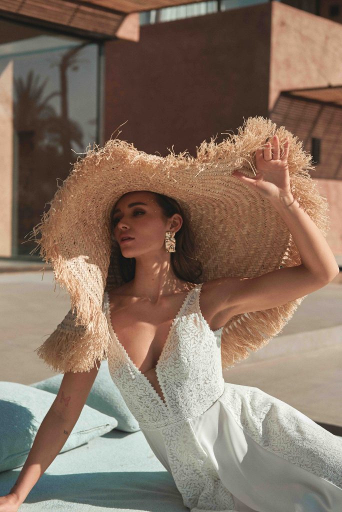 In questa immagine un abito da sposa della collezione Adore by Justin Alexander 2024 con corpetto in pizzo e maxi cappello di paglia. 