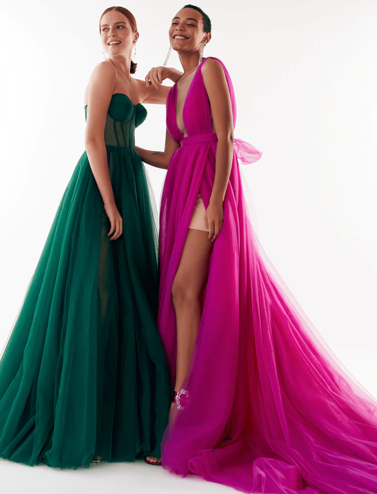 In questa foto due modelle posano con gli abiti da cerimonia donna 2024 di Millanova: si tratta di due vestiti voluminosi, in tulle, nei toni del verde bosco e del fucsia