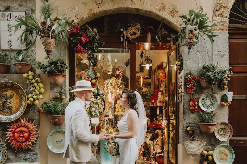 In questa foto due sposi di un Elopement Wedding di Tania Costantino davanti ad un  negozio tipico di Cefalù