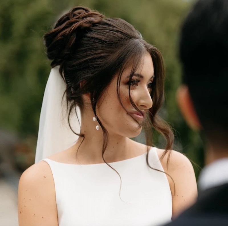 In questa foto una sposa con un'acconciatura con chignon molto alto e ciocche di capelli attorno al viso