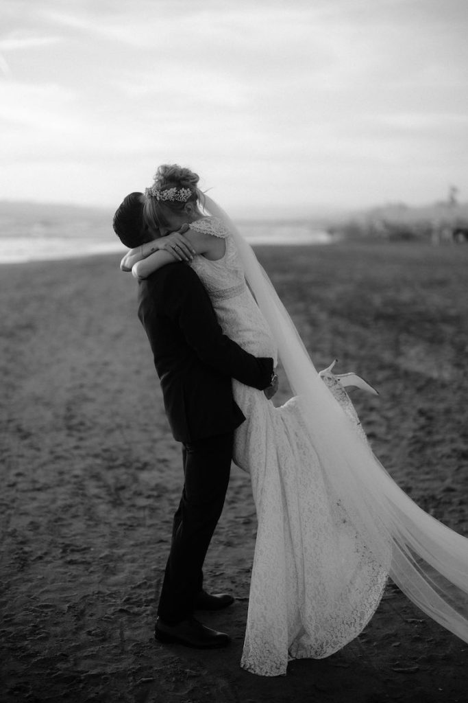 In questa foto in bianco e nero di Alessio Bazzichi due sposi abbraccia su una spiaggia. Lo sposo la solleva leggermente da terra