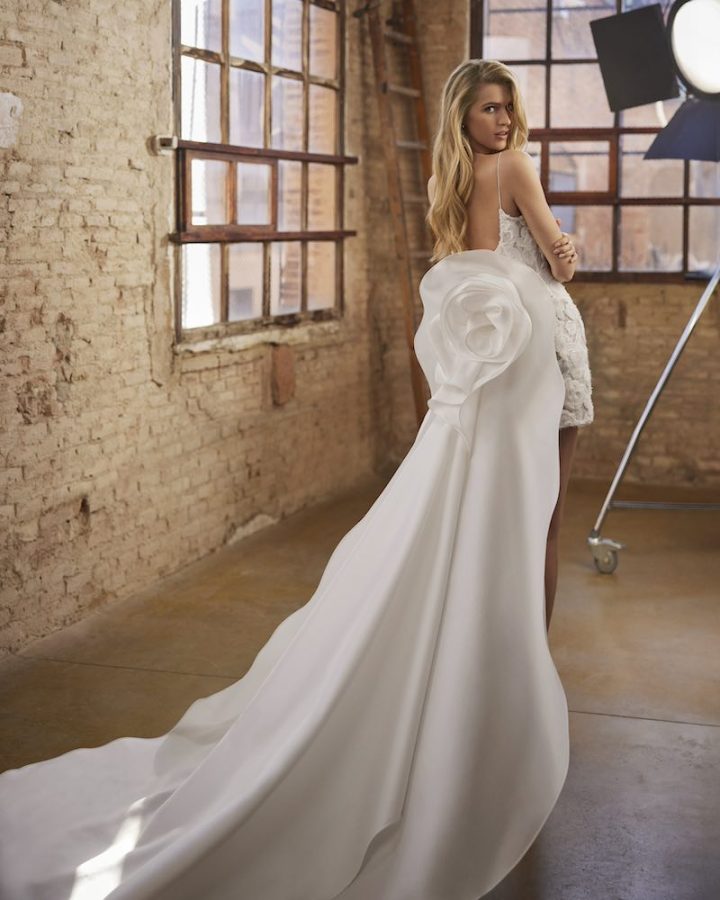 In questa foto una modella posa di spalle indossando un abito da sposa Aire Atelier 2025 corto con sovragonna staccabile decorata da un maxi fiore 3D