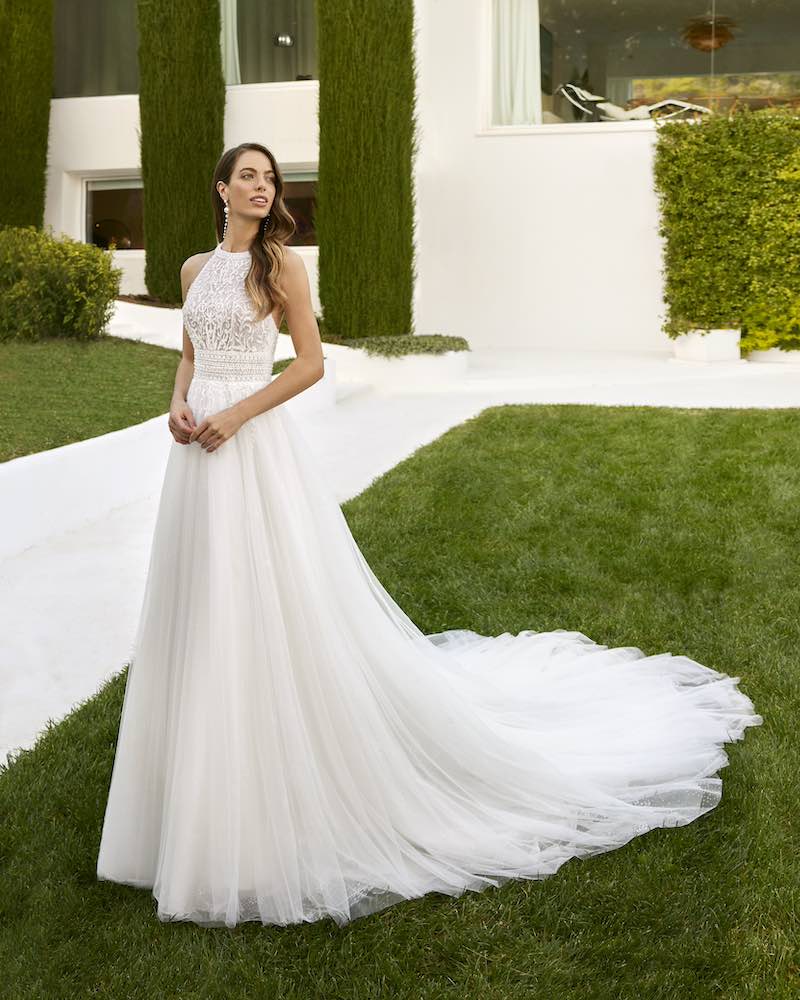 In questa foto una modella posa indossando un abito da sposa Aire Chic 2025 con corpetto i pizzo con scollo all'americana e gonna ad A in tulle