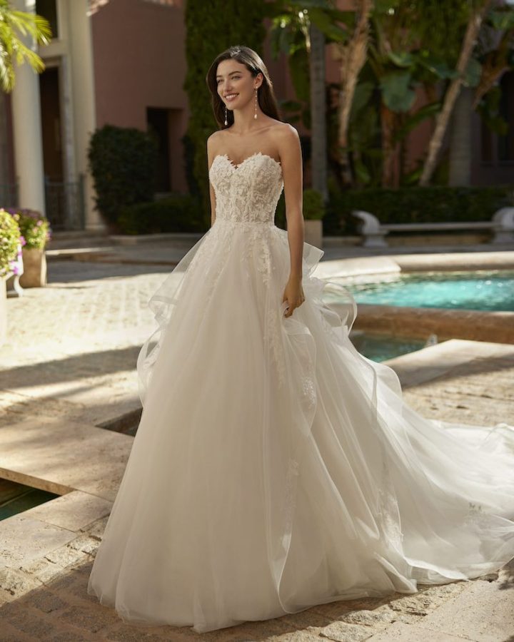 In questa foto una modella posa indossando un abito da sposa Aire Royale 2025 con gonna ad A in tulle e corpetto in pizzo floreale con scollo a cuore