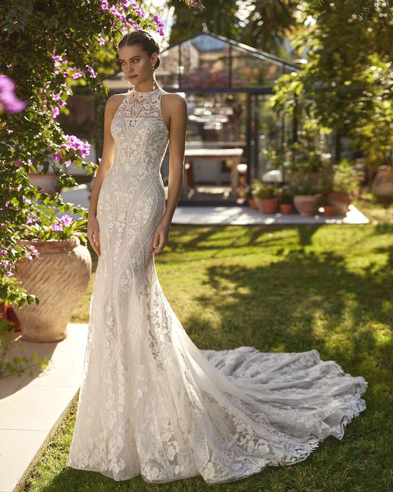 In questa foto una modella posa indossando un abito da sposa Aire Boho 2025 a sirena in pizzo con scollatura all'americana