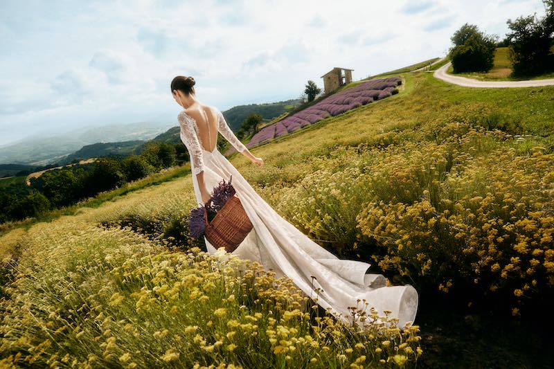 In questa foto la modella Luma Grothe posa di spalle in un campo di elicriso e lavanda indossando un abito ad A della nuova collezione sposa Atelier Emé 2025