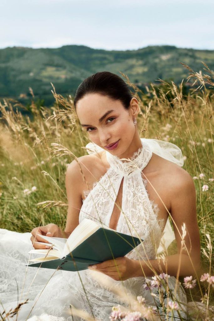 In questa foto la modella Luma Grothe posa indossando un abito della collezione sposa Atelier Emé 2025, mentre legge un libro seduta su un prato