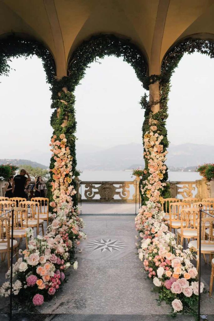 In questa foto la Loggia Durini di Villa del Balbianello allestita da Imène Hanafi per la cerimonia del matrimonio di Carrie e Adam con fiori nei toni del rosa e sedie di colore dorato 