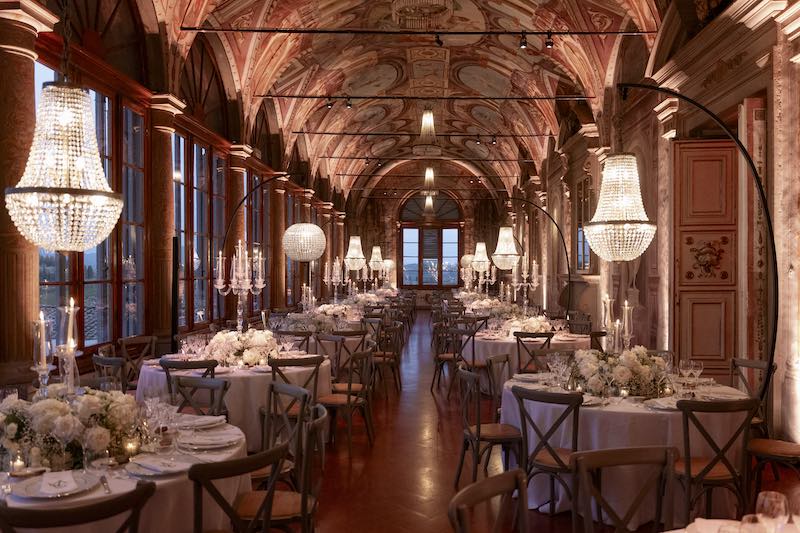 In questa foto, la Galleria di Villa Corsini a Mezzomonte allestita per un matrimonio con lampadari, tavoli rotondi, fiori bianchi e tovagliato en pendant