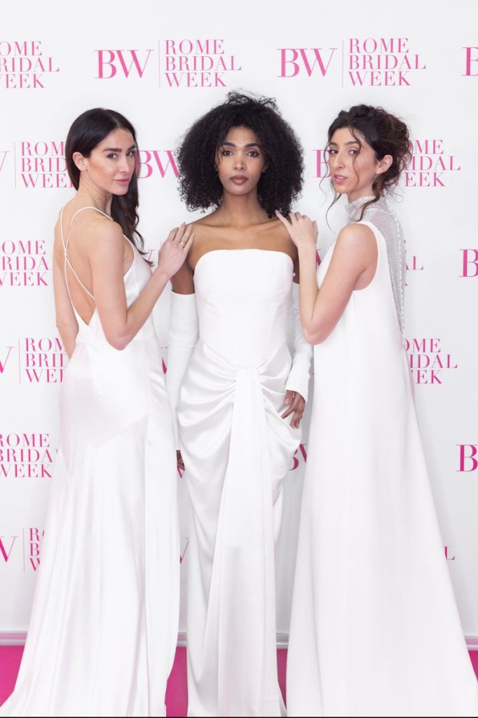 In questa foto, tre modelle in abiti da sposa posano davanti al roll-up della Rome Bridal Week