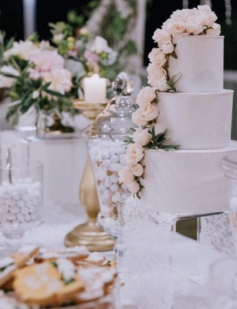 In questa foto uno sweet table decorato nei toni del bianco e del rosa cipria. Sul tavolo a piani sono disposti la wedding cake deocrata con rose rosa e biscotti di pasta frolla