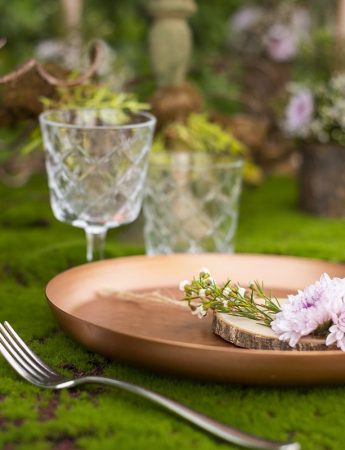 In questa foto una mise en place realizzata con muschio perfetta per un matrimonio ecochic. Sui piatti colore gold rose sono disposte piccole margherite rosa. I centrotavola sono campane di vetro che racchiudono fiori in coordinato